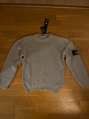 Ljusgrå stickad tröja från Stone Island - Säljer en ljusgrå stickad tröja från Stone Island med klassisk patch på ärmen. Tröjan har rund halsringning, ribbade muddar och är långärmad. Perfekt för dig som gillar stilrena och tidlösa plagg.