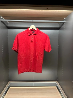 Röd pikétröja från Tommy Hilfiger - Klassisk röd pikétröja från Tommy Hilfiger med liten logga på bröstet och två knappar vid kragen. Tröjan har korta ärmar och är gjord i mjuk bomull. Perfekt till jeans eller shorts för en clean och enkel look.