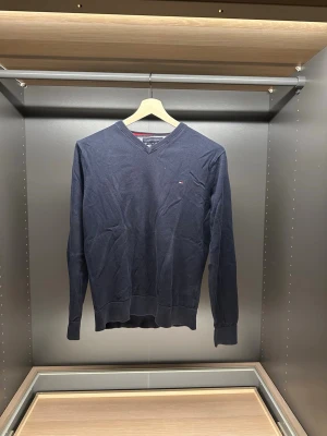 Mörkblå V-ringad tröja Tommy Hilfiger - Klassisk mörkblå V-ringad tröja från Tommy Hilfiger i premium bomull. Diskret logga på bröstet och ribbade muddar vid ärmslut och nederkant. Perfekt till lager på lager och enkel att matcha med jeans eller chinos.