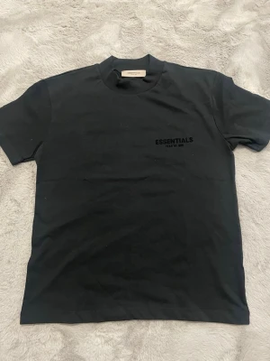 Svart Essentials Fear of God t-shirt - Säljer en svart t-shirt från Essentials Fear of God med tryck både fram och bak. Klassisk passform med rund hals och korta ärmar. T-shirten är gjord i mjuk bomull och har en clean streetwear-look med diskret logga framtill och stor logga på ryggen.