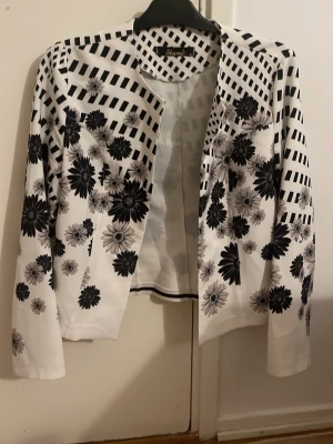 Vit och svart blommig kavaj från Shamo - Säljer en unik kavaj från Shamo i vitt med svart blommönster och rutiga detaljer. Kavajen har lång ärm, öppen framtill och är tillverkad i ett lätt, syntetiskt material. Perfekt för dig som vill sticka ut med en cool och modern look.