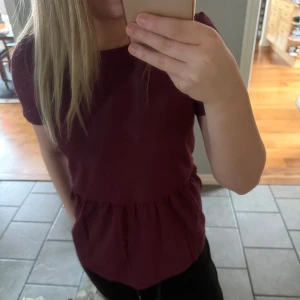 Vinröd topp från Lindex - Säljer en vinröd topp från Lindex i mjuk bomull. Modellen har rund halsringning, korta ärmar och ett rynkat parti i midjan som ger en söt siluett. Perfekt till jeans eller kjol för en enkel och snygg look.