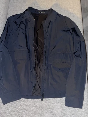 Zara - Säljer en stilren mörkblå vindjacka från Zara. Jackan har klassisk krage, två stora fickor framtill och stängs med dragkedja. Perfekt för dig som vill ha en enkel och lätt jacka till vardags. Materialet är polyester och jackan har lång ärm.