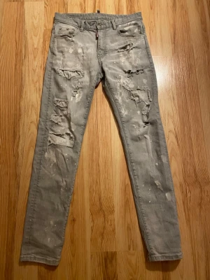Dsquared2 ljusgrå jeans - Säljer ett par grå jeans från Dsquared2 med slitna detaljer och färgstänk för en riktigt edgy look. Byxorna har normal passform, fem fickor och klassisk knappgylf. Perfekta för dig som vill sticka ut med en streetwear vibe.