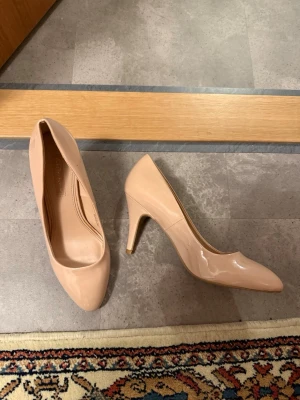 Rosa pumps med klack - Säljer ett par stilrena pumps i rosa lack med spetsig tå och hög klack. Skorna har en klassisk form och är tillverkade i syntetmaterial med glansig finish. Perfekta för dig som vill ha en clean och elegant look.
