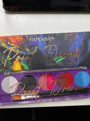 Handaiyan Paint Dream ögonskuggspalett - Färgstark ögonskuggspalett från Handaiyan med fem intensiva nyanser: vit, svart, rosa, röd och blå. Paletten har genomskinligt lock med guldtext och medföljande pensel. Perfekt för kreativa sminkningar och unika looks.