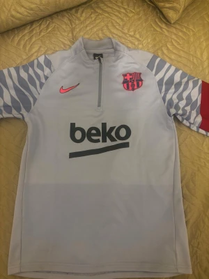 Barça Nike träningströja halvzip - Säljer en ljusgrå FC Barcelona träningströja från Nike med halv dragkedja. Tröjan har zebramönstrade ärmar i grått och vitt, rosa Nike-logga och Barça-emblem samt svart 'beko'-tryck på bröstet. Perfekt för dig som älskar fotboll och vill ha en snygg och bekväm träningsstil.