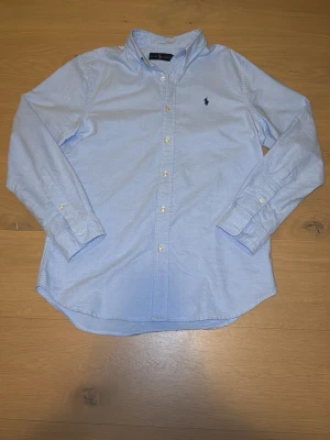 Skjorta från Ralph Lauren - Klassisk ljusblå skjorta från Ralph Lauren med button-down krage och broderad logga på bröstet. Skjortan har långa ärmar, vita knappar och är gjord i mjuk bomull. Perfekt till jeans eller chinos för en clean look.