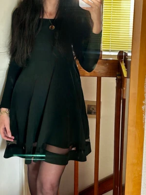 Svart kort klänning med meshdetalj - Säljer en svart kort klänning med lång ärm och rund halsringning. Kjolen har en lätt utställd form och en snygg meshdetalj längst ner som ger en modern touch. Perfekt för dig som gillar stilrena plagg med det lilla extra.