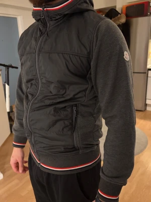 Svart pufferjacka från Moncler - Snygg svart pufferjacka från Moncler med huva och dragkedja. Jackan har stickade ärmar och ribbade muddar med röd, vit och blå rand. Perfekt för dig som vill ha en sportig och stilren look. Moncler-logga på ärmen och praktiska fickor med dragkedja.