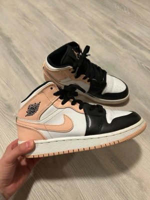 Nike Air Jordan 1 Mid Peach/Black - Nike Air Jordan 1 Mid sneakers i en snygg färgkombo med persikofärgade, svarta och vita paneler. Skorna har klassisk snörning, rund tå och platt sula. Materialet är skinn med detaljer i syntet. Ikonisk Jumpman-logga på plösen och Air Jordan-märke på sidan.