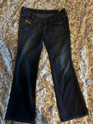 Mörkblå bootcut jeans från diesel - Snygga mörkblå jeans från Diesel med bootcut-modell och coola broderade detaljer på fickorna. Klassisk femficksdesign och kontrastsömmar. Jeansen är i stretchigt denim och har en låg midja för en trendig look.
