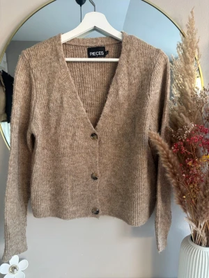 Beige stickad kofta från Pieces - Mysig beige stickad kofta från Pieces i storlek S. Koftan har v-ringning, tre knappar framtill och långa ärmar. Perfekt för lager på lager och ger en avslappnad men ändå snygg look.