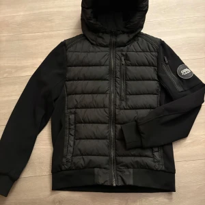 Svart jacka Supply & Demand - Säljer en svart jacka i storlek S från Supply & Demand med huva och quiltad framsida. Jackan har dragkedja framtill, fickor med dragkedja och en patch på ärmen. Materialmix med polyester och syntet, perfekt för kyligare dagar. Snygg och modern look.