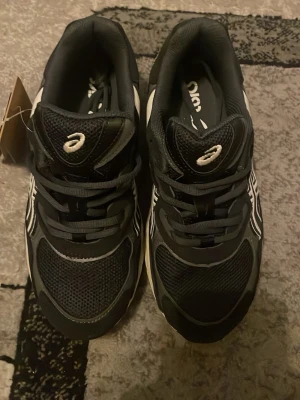 Svarta Asics sneakers med mesh och skinn - Säljer ett par soprena svarta Asics sneakers med vita detaljer. Skorna har en chunky sula, meshpartier för extra andningsförmåga och klassisk snörning. Perfekta för dig som vill ha både stil och komfort i vardagen.