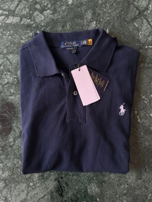 Marinblå pikétröja från Polo Ralph Lauren - Helt ny Klassisk marinblå pikétröja från Polo Ralph Lauren med vit broderad logga på bröstet. Tröjan har krage, korta ärmar och knäppning framtill. Tillverkad i mjuk bomull som känns skön mot huden. Perfekt för en clean och stilren look. Hör gärna av dig vid funderingar🤩