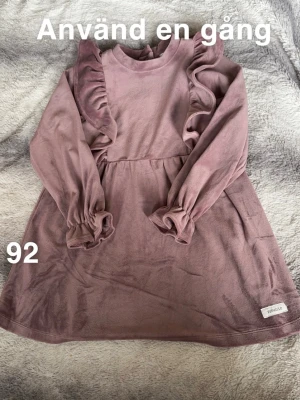 lila klänning med volang från Newbie - Supersöt lila klänning från Newbie. Klänningen har långa ärmar med volangdetaljer längs axlarna och resår vid ärmsluten för extra söt look. Perfekt för dig som gillar gulliga och bekväma plagg.🌸Använd en gång. Storlek 92 