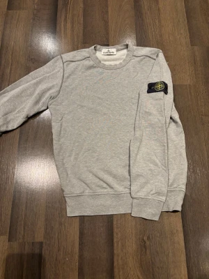 Stone Island Junior - Säljer en stilren grå sweatshirt från Stone Island Junior med klassisk patch på vänster ärm. Tröjan har rund hals, ribbade muddar och är tillverkad i mjuk bomull. Perfekt för dig som gillar enkel och clean streetwear.