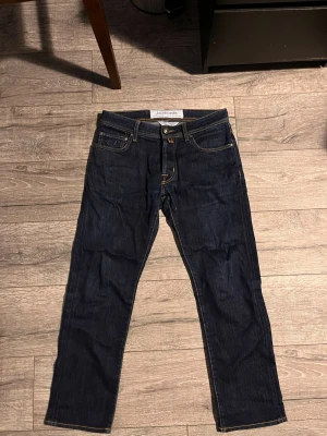 Mörkblå jeans från Jacob Cohen - Säljer ett par mörkblå jeans från Jacob Cohen med klassisk femficksdesign och kontrastsömmar. Jeansen har en snygg blå patch bak i midjan och raka ben. Perfekta för en stilren och avslappnad look. Storlek 30 skulle säga att dom är lite mindre i storleken och passar dig som är 170-175