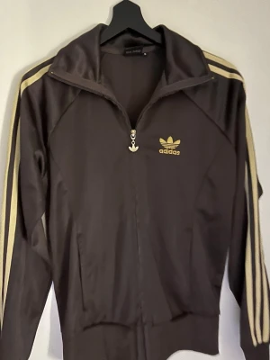 Brun Adidas ziptröja med guldiga ränder - Säljer en brun Adidas ziptröja i storlek S med klassiska guldiga ränder längs ärmarna och Adidas-logga på bröstet. Tröjan har dragkedja hela vägen. 