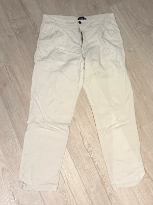 Beiga chinos från Lager - Snygga beiga chinos från 157 med klassisk passform och knappstängning. Byxorna har framfickor och är tillverkade i mjuk bomull som känns skön mot huden. Perfekta för dig som gillar en clean och enkel stil.