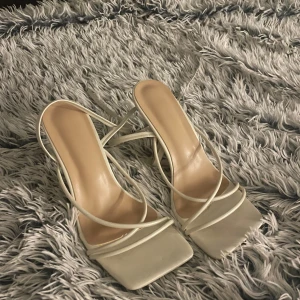 Vita sandaler med smala remmar och klack - Säljer ett par trendiga vita sandaler med smala remmar och fyrkantig tå. Skorna har hög klack och öppen tå, vilket ger en stilren och modern look. Tillverkade i syntetmaterial med justerbar rem runt ankeln. Perfekta för dig som vill ha en clean och chic vibe. Har bara använd dem en dag på en födelsedagsfest 