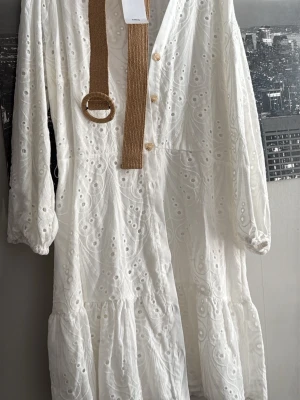 Vit broderad långklänning Mango - Superfin vit långklänning från Mango med broderade detaljer och knappar framtill. Klänningen har långa ärmar med resår vid ärmslut och medföljer ett beige bälte i flätad stil. Perfekt för sommarens alla tillfällen!
