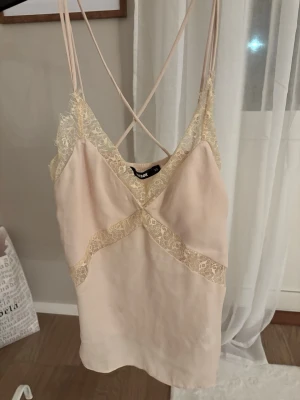 Beige linnetopp med spets från Bik Bok - Säljer en superfin beige linnetopp från Bik Bok med tunna axelband som korsas i ryggen. Toppen har vackra spetsdetaljer längs urringningen och tvärs över framsidan. Perfekt för sommaren och ger en romantisk vibe.