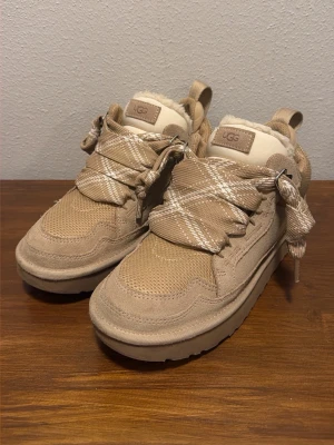 UGG lowmel trainer ”sand” - Säljer ett par beige sneakers från UGG med breda snören och vitrutigt mönster. Skorna har en rund tå, platt sula och är fodrade med mjuk päls för extra värme. Materialmix av mocka och syntet, perfekta för kyligare dagar.