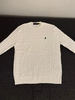 Vit kabelstickad tröja Polo Ralph Lauren - Vit kabelstickad tröja från Polo Ralph Lauren med klassisk rund halsringning och broderad marinblå logga på bröstet. Tröjan har lång ärm och ribbade muddar vid ärmslut och nederkant. Perfekt för lager-på-lager under kyliga dagar.