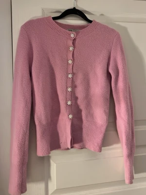 Rosa kofta med pärlknappar från Zara - Supersöt rosa kofta från Zara med långa ärmar och fina pärlformade blomknappar framtill. Koftan är stickad och har en figurnära passform som ger en feminin look. Perfekt att styla med jeans eller kjol för en trendig vibe.