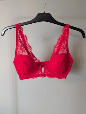 Röd spets-bh - Snygg röd bh i spets med dekorativ rosett framtill och transparenta detaljer. Axelbanden är justerbara och kuporna har en lätt vaddering. Spetsen har ett blommigt mönster och bh:n har en liten cut-out mellan kuporna för extra detalj.