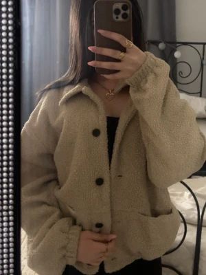 Beige teddyjacka med stora knappar - Supermjuk och fluffig beige teddyjacka med stora svarta knappar framtill och två rymliga fickor. Jackan har en avslappnad passform och långa ärmar med resår vid handlederna. Perfekt för dig som vill ha en cozy look under kyliga dagar.