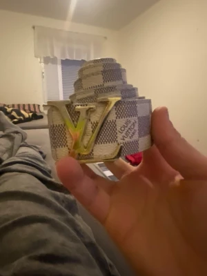 Louis Vuitton rutig bälte damier - Säljer ett Louis Vuitton bälte med klassiskt damiermönster i beige och grå toner. Bältet har ett stort LV-spänne i guld och är tillverkat i ett tåligt syntetmaterial. Perfekt accessoar för att lyfta din outfit med en lyxig touch.