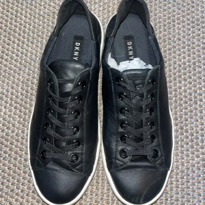 Svarta sneakers från DKNY - Snygga svarta sneakers från DKNY med vit platt sula och tydlig logga runt hela sulan. Skorna har rund tå, snörning och är gjorda i skinn. Perfekt för dig som gillar stilrena och trendiga skor med tydlig branding.