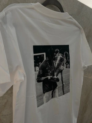 Ronaldo T-shirt - Lyft din stil med vår vita premium T-shirt med ett svartvitt tryck av Ronaldo på ryggen. En stilren och kraftfull design som hyllar en av världens största fotbollsstjärnor – perfekt för både vardag och streetwear-look. T-shirten är tillverkad i mjuk och slitstark bomull som ger hög komfort hela dagen. Den minimalistiska framsidan kombineras med ett iögonfallande motiv på ryggen, vilket skapar en modern och trendig kontrast  