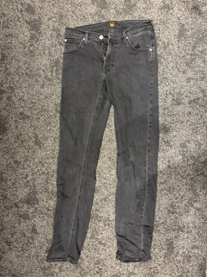 Svarta Lee jeans - Säljer ett par svarta jeans från Lee med klassisk femficksdesign och dragkedja. Jeansen har en smal, rak passform och är tillverkade i mjukt denimtyg. Snygga detaljer med Lee-logga på bakfickan och läderpatch i midjan. Perfekta till sneakers eller boots.