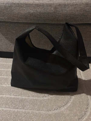 Svart axelväska i skinnimitation - Stilren svart axelväska med enkel design och mjuk struktur. Väskan har ett brett axelband och är tillverkad i skinnimitation med lätt kornig yta. Perfekt storlek för att få plats med allt du behöver till skolan eller stan.