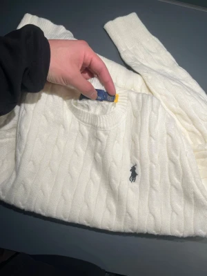 Vit kabelstickad tröja Polo Ralph Lauren - Kabelstickad vit tröja från Polo Ralph Lauren med klassisk rund hals och broderad marinblå logga på bröstet. Tröjan är långärmad och har ribbade muddar vid ärmslut och nederkant. Perfekt för dig som gillar stilrena och tidlösa plagg.