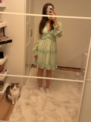 Grön blommig kortklänning Vero Moda - Supersöt ljusgrön kortklänning från Vero Moda med blommigt mönster och volangdetaljer. Klänningen har v-ringning, långärmade transparenta ärmar och omlott-effekt upptill. Materialet är lätt och flowy, perfekt för varma dagar. Passar dig som gillar romantisk stil!