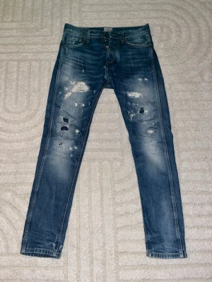 Blå slitna jeans från Jack & Jones - Säljer ett par vintage blå jeans från Jack & Jones med snygga slitningar och hål på framsidan. Modellen är slim fit med normal passform och klassisk femficksdesign. Jeansen har en mörk tvätt med ljusare partier och är gjorda i bomullsmaterial. Modellen heter Erik anti fit. Storleken är avklippt, men skulle säga att det är w28-29 och längden är L 30. Modellen är 175 cm lång och väger 59kg 