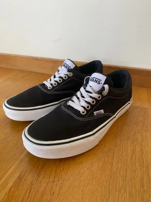 Svarta Vans sneakers i storlek 40 - Klassiska svarta Vans sneakers med vit sula och vita snören. Skorna har låg profil, rund tå och tydliga kontrastsömmar. Tillverkade i slitstark canvas med Vans-logga på sidan. Säljes eftersom de är för små nu. Skorna är i storlek 40 och i fint skick. Hör gärna av er vid eventuella frågor.
