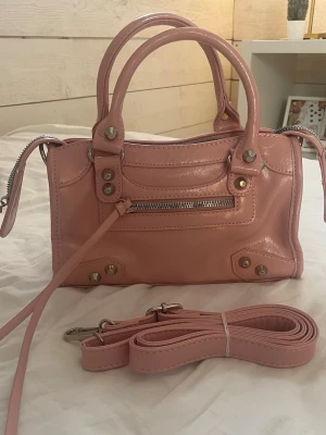 Handväska rosa - Säljer en snygg rosa handväska i skinn med silverfärgade detaljer och dragkedja framtill. Väskan har både handtag och en avtagbar axelrem, samt ett rymligt innerfack och en mindre ficka med dragkedja. Perfekt för dig som vill ha en stilren och trendig väska. Jag väljer att sälja den då den inte kommer till användning.