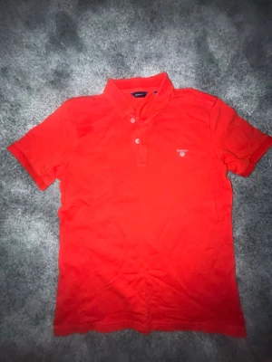 Röd pikétröja från GANT, 13-14 år - Snygg röd pikétröja från GANT med klassisk krage och knappar framtill. Tröjan har broderad GANT-logga på bröstet och är gjord i 100% bomull för en skön känsla. Perfekt till jeans eller shorts för en clean look.