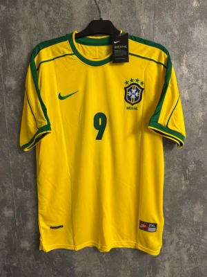 Brasilien Ronaldo - Retro Ny storlek 2XL - Bilderna visar en gul Brasilien fotbollströja från Nike med gröna detaljer och nummer 9, samt namnet Ronaldo på ryggen. Tröjan är i storlek 2XL och tillverkad i Thailand med Nike Breathe-teknologi.