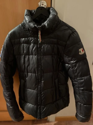 Svart pufferjacka från Dolomite - Svart pufferjacka från Dolomite med quiltad design och hög krage. Jackan har en avtagbar huva med beige pälsdetalj och dragkedja framtill. Märkespatch på ärmen och snygga detaljer vid kragen. Perfekt för kalla dagar och riktigt stilren.