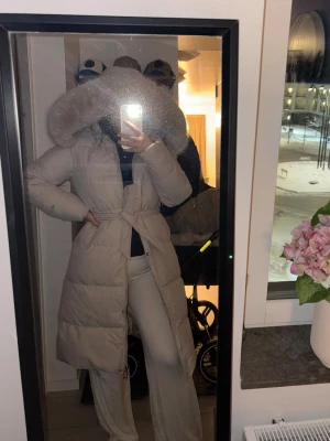 Beige lång dunjacka med päls - Säljer en beige lång dunjacka med fluffig päls runt huvan. Jackan har bälte i midjan, dragkedja och knappar framtill. Den är quiltad och har långa ärmar, perfekt för kalla vinterdagar. Snygg och varm stil som sticker ut med den stora pälsdetaljen. Knappt använd. En av knappars med ena sidan har gått av ( dock inget som syns) 