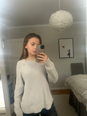 Ljusgrå stickad tröja med knappar - Säljer en ljusgrå stickad tröja med rund hals och snygga knappar längs axeln. Tröjan har lång ärm, slits i sidorna och en relaxed passform. Perfekt för lager-på-lager och chill dagar.