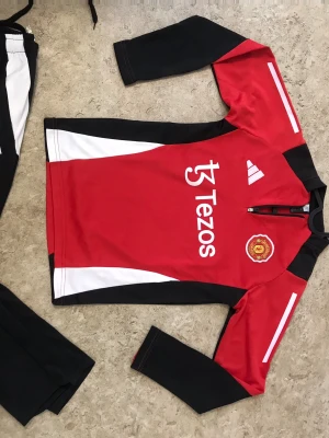 Manchester United träningströja röd/svart - Säljer en Manchester United träningströja från Adidas i rött, svart och vitt. Tröjan har lång ärm, halv dragkedja och klubbmärke på bröstet. Materialet är polyester och designen har vita detaljer och logotypen Tezos tryckt framtill.