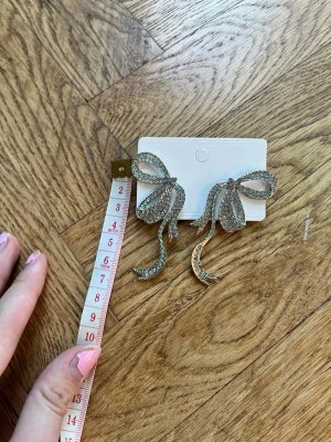 Glittriga rosettörhängen i silver - Säljer ett par stora örhängen i form av rosetter, täckta med glittrande stenar. De är silvriga och riktigt statement, perfekta för att lyfta vilken outfit som helst. Supersnygga till både uppsatt och utsläppt hår!